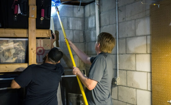two pest control technicians cleaning spider webs from a basement ceiling using a telescoping brush